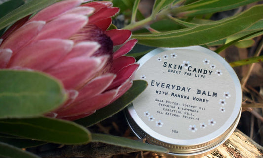 Tin of 'Skin Candy' Everyday Balm with a pink flower and green leaves in the background